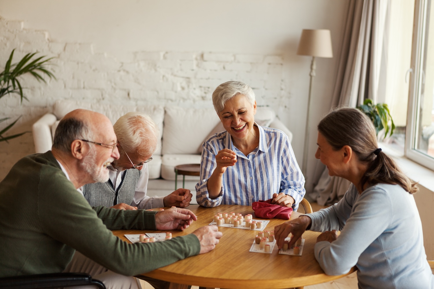Choosing a Nursing Home in Bloomfield Hills A group of four older adults sitting around a table, smiling and laughing while playing a board game together in a bright, cozy living room.