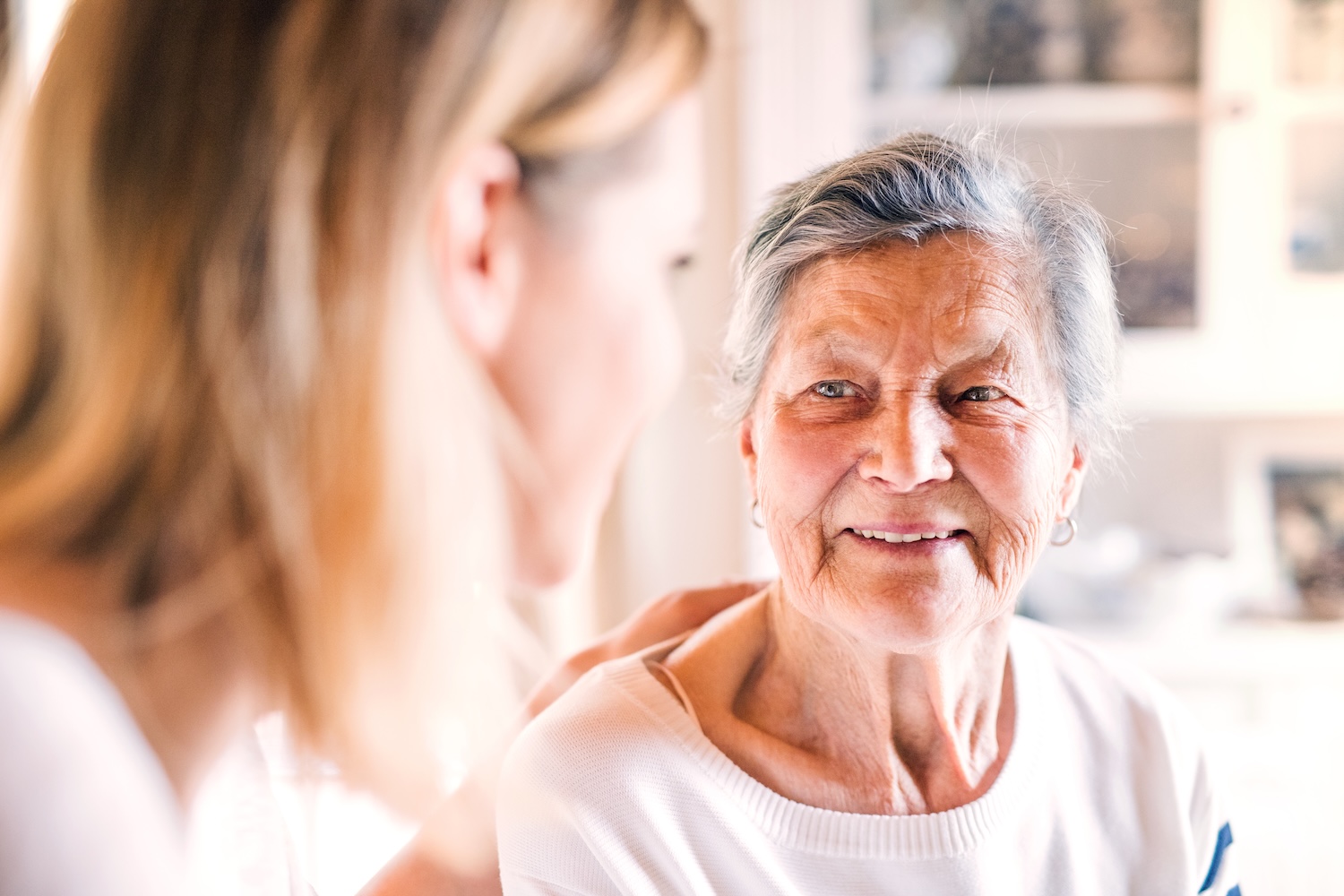 How to Move a Parent With Dementia to Assisted Living A Helpful Guide Elderly woman smiling warmly at a younger woman who is sitting beside her, gently resting a hand on her shoulder, sharing a moment of connection in a bright, cozy room.